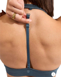 Back view of a person adjusting the removable center-back strap of the ORU HALTER SPORTS BRA in blue with light to medium support and a sleek inverted V hemline.